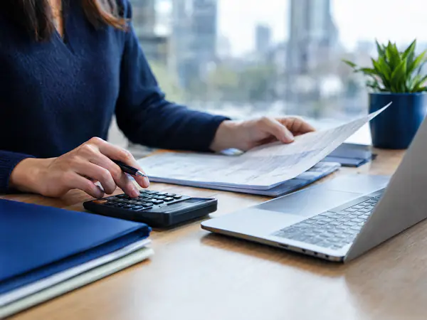 Hands reconciling bank statements with a calculator and laptop beside neatly stacked folders
